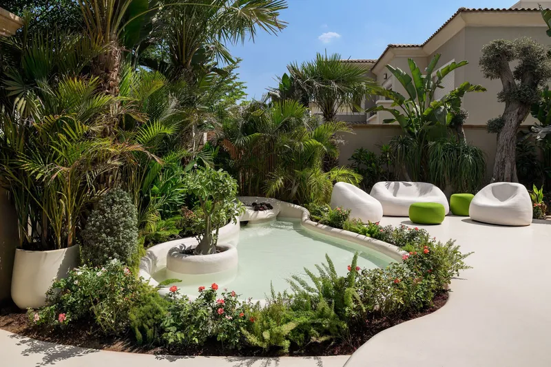 Outdoor courtyard with a shallow pool, planted beds, palms, and seating under blue sky