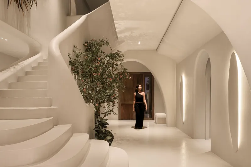 White curved entry hall with spiral staircase and arched doorways, a flowering tree and a visitor in evening light