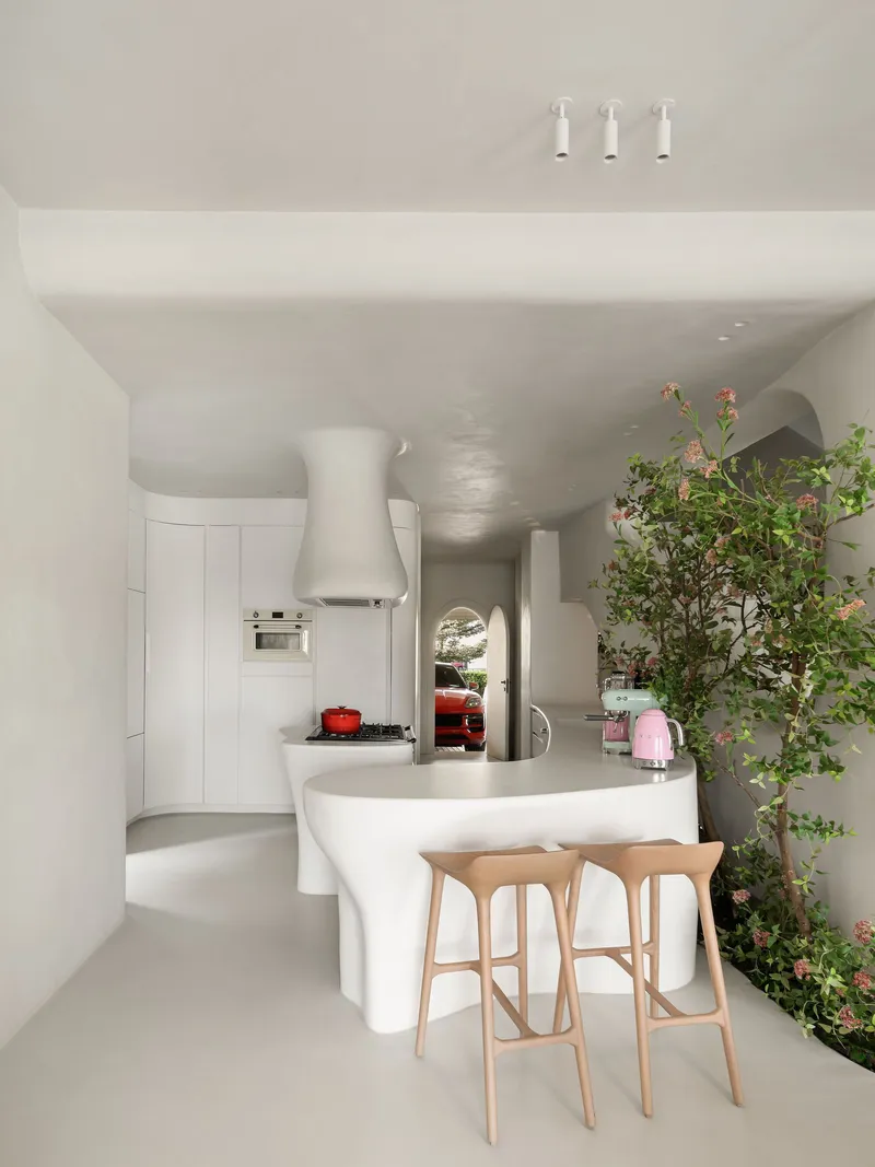 Open kitchen with curved white island, integrated range hood and flowering tree beside timber bar seating