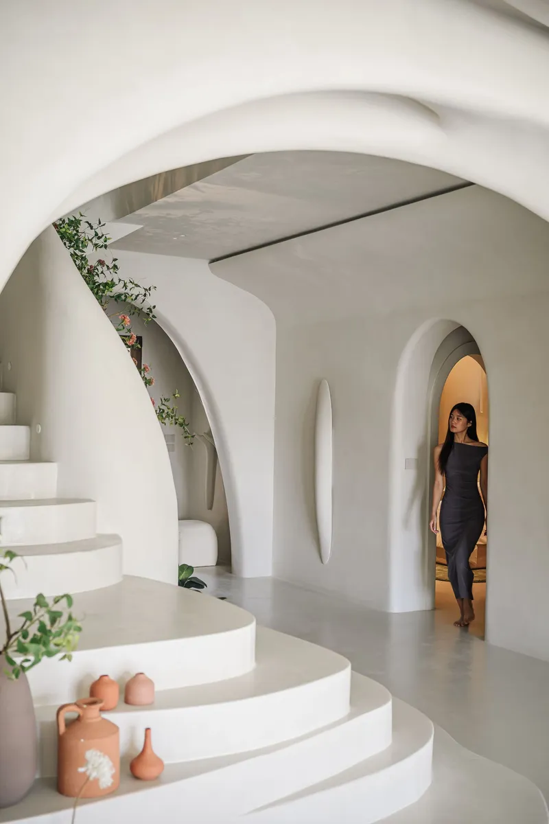 White curved staircase with integrated planters and arched doorway where a woman stands in afternoon light