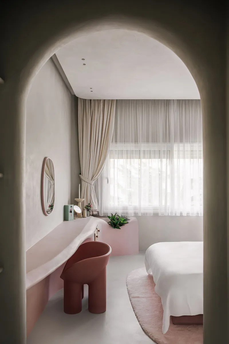 Bedroom viewed through rounded archway with pink ledge desk and terracotta chair facing draped windows