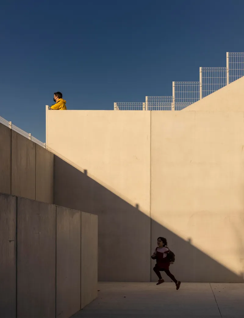 Concrete walls and courtyard with child running across sunlit pavement and figure above on parapet