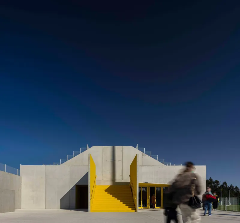 Concrete facade with central yellow metal staircase and blurred figures at entrance under blue sky