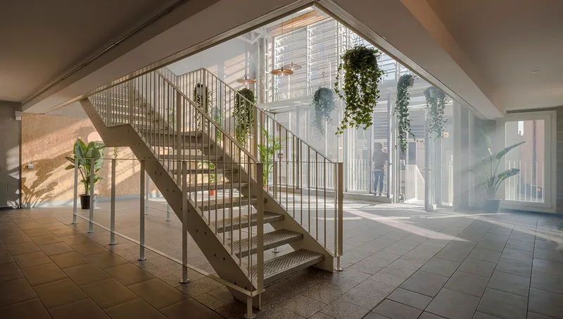 Metal staircase with vertical rod railings rising through a skylit lobby with potted plants and sunlight