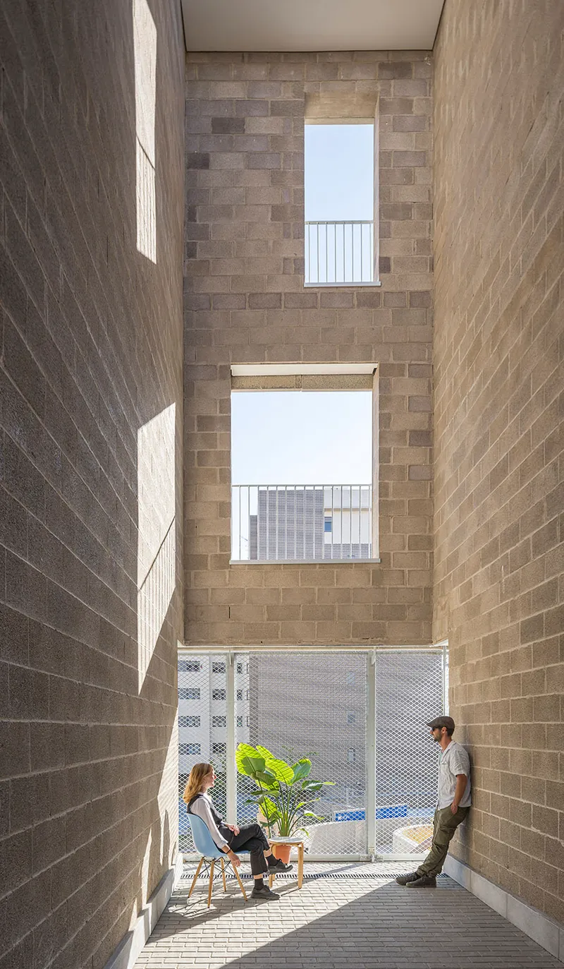 Narrow courtyard with brick walls, stacked window openings, and two people seated near a potted plant