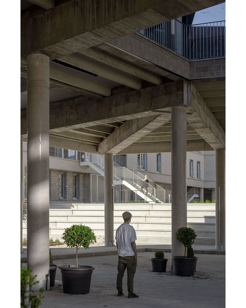 Covered courtyard with cylindrical concrete columns and a person standing among potted trees