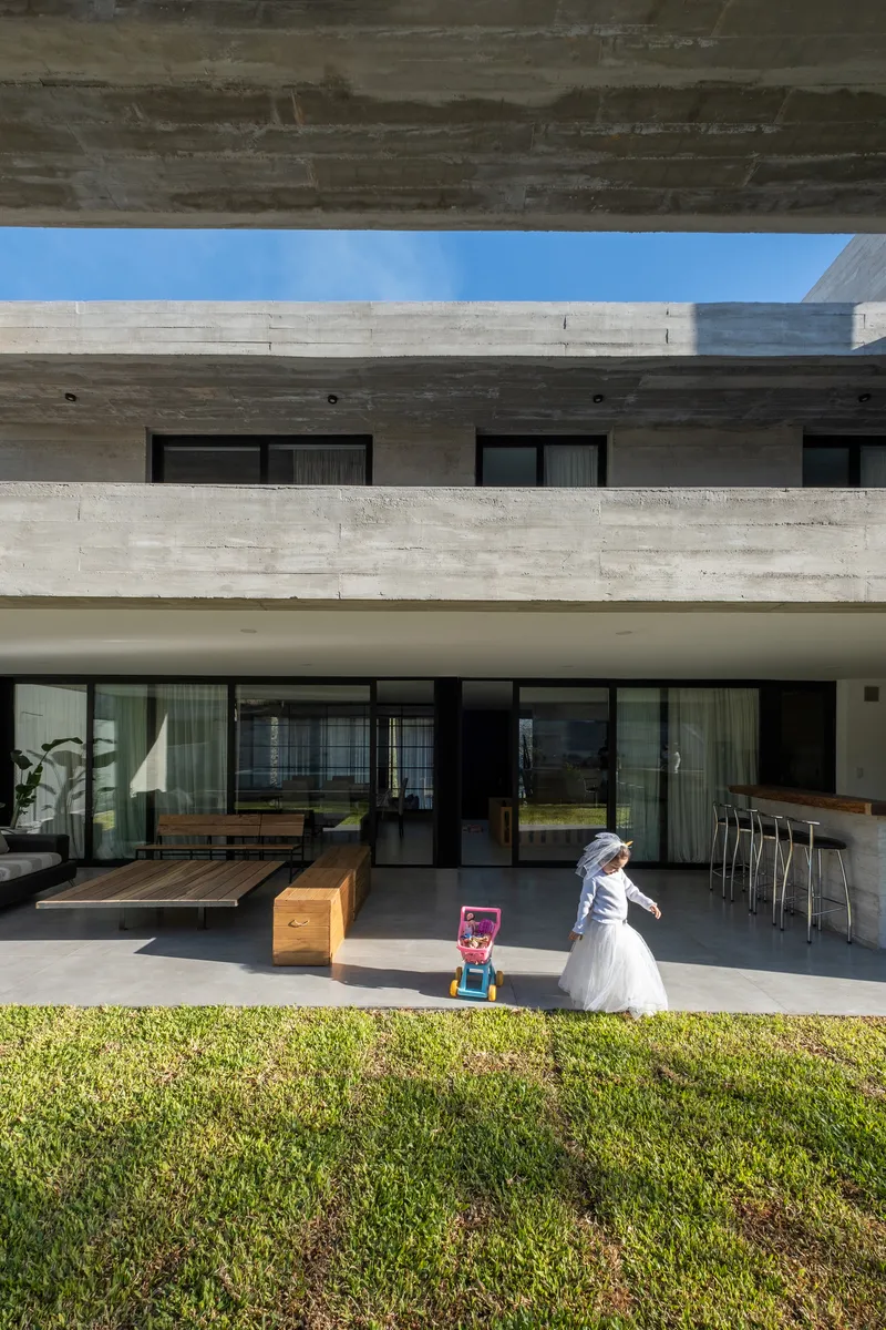 Covered terrace with board-formed concrete beams and a child playing on the lawn in morning light