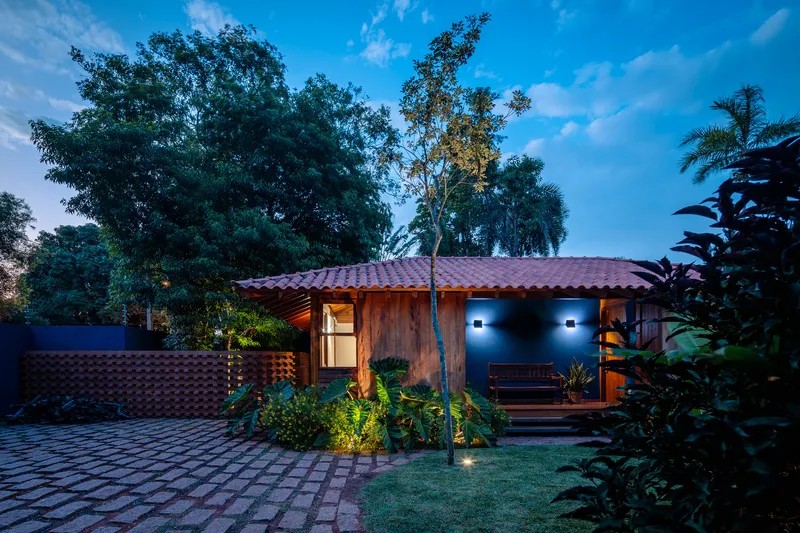 Timber-clad entrance facade with terracotta tile roof illuminated at dusk among garden trees