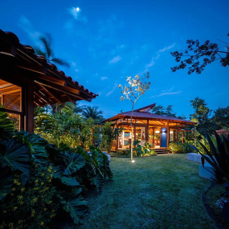 Garden view of timber pavilions with clay tile roofs surrounded by tropical plantings at dusk