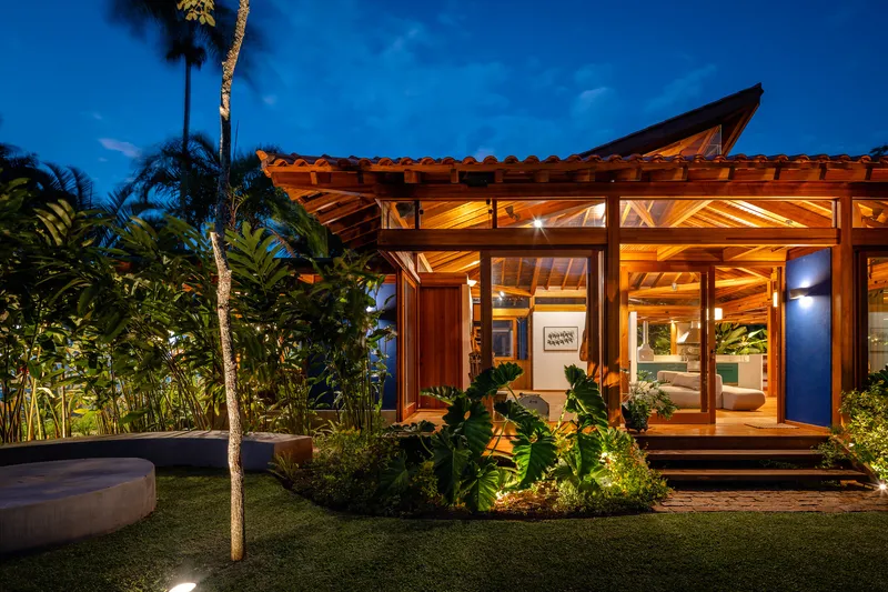 Glazed facade with exposed timber trusses and overhanging eave framed by garden palms at twilight