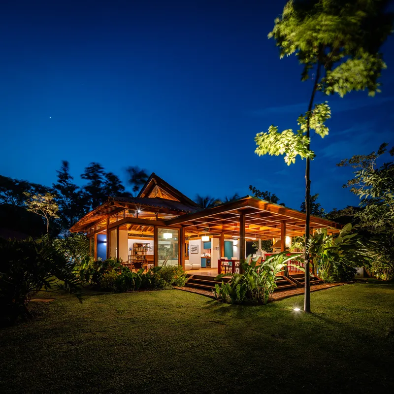 Single-story pavilion with pyramidal roof and timber pergola viewed across lawn under evening sky