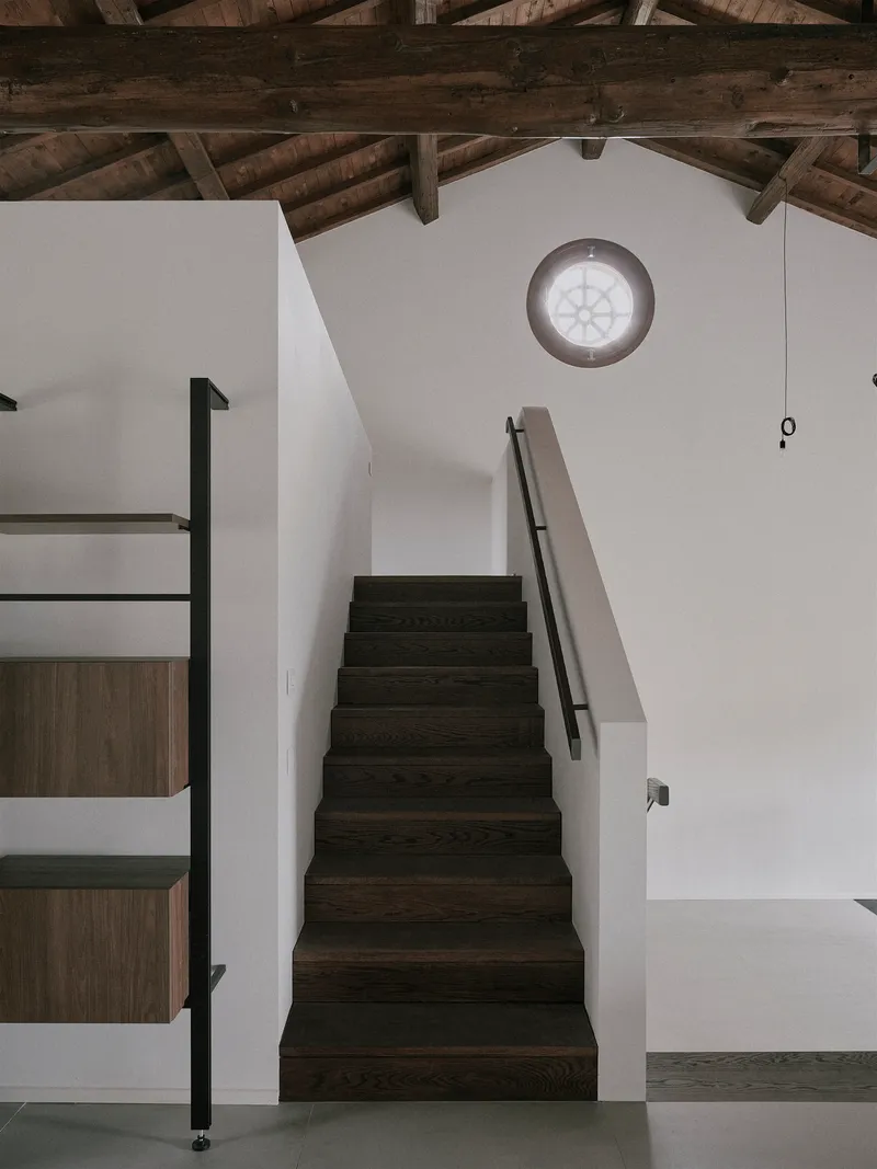 Dark timber staircase with metal handrail rising beneath exposed roof beams and a circular window