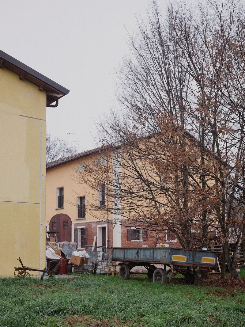Yellow-rendered building volumes with bare deciduous trees and farm trailer in overcast weather
