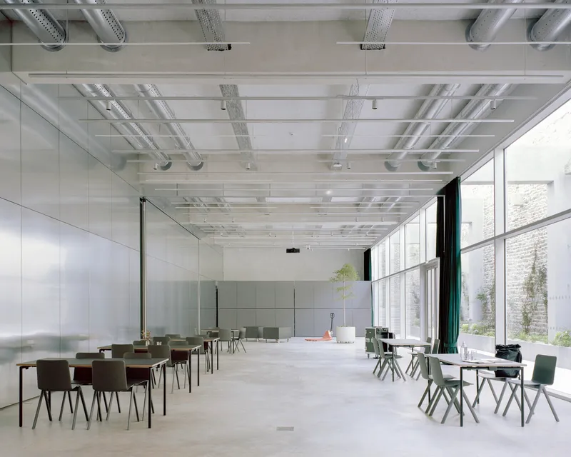 Multipurpose room with folding stainless steel partitions, concrete floor, and floor-to-ceiling windows overlooking a courtyard
