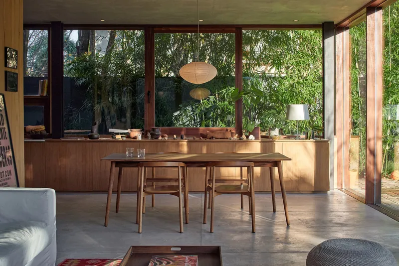 Open plan dining area with timber table and floor-to-ceiling glazing overlooking planted vegetation