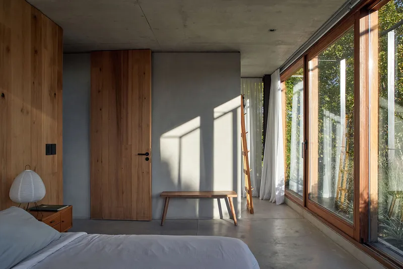 Bedroom with exposed concrete ceiling and walls, timber-framed windows casting angular shadows across polished floor