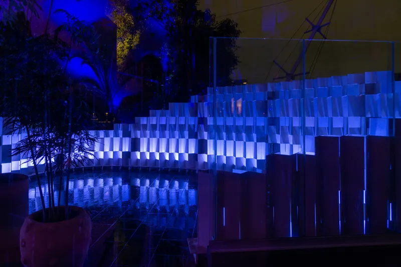 Illuminated panel wall reflected in a dark water feature with planted trees nearby