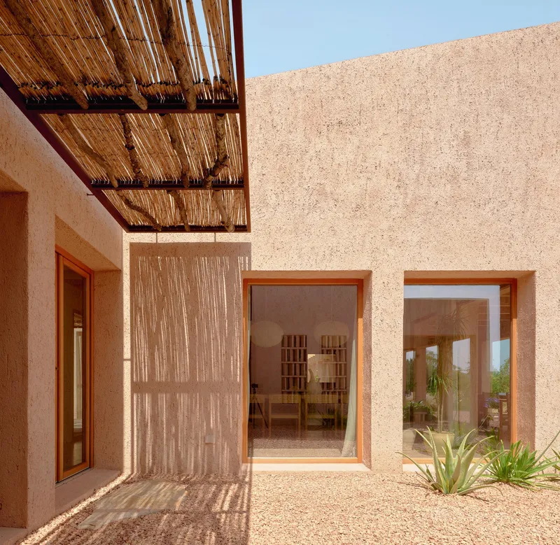 Courtyard with textured aggregate walls, bamboo overhead canopy casting shadows, and glazed doors opening to interior spaces