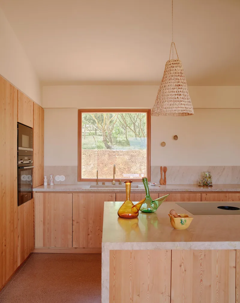 Kitchen with timber cabinetry, marble countertops, woven pendant light, and window framing view of olive trees