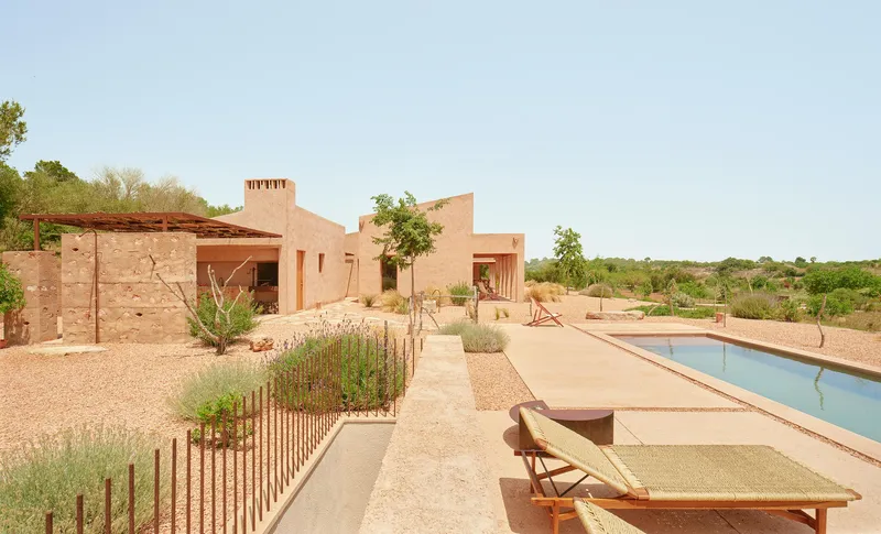 Rammed earth compound with pool terrace, planted beds and loungers under afternoon light