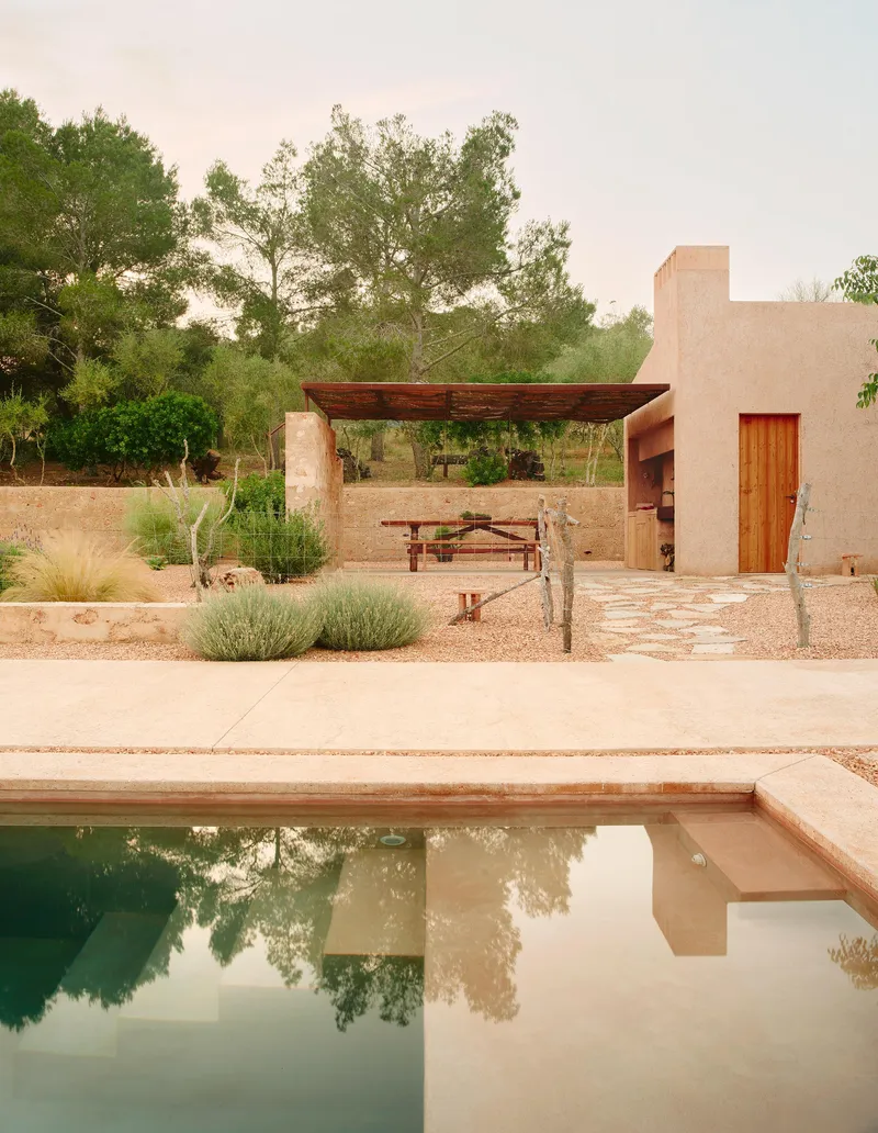 Pool reflecting straw-textured walls and timber pergola structure in planted courtyard under overcast sky