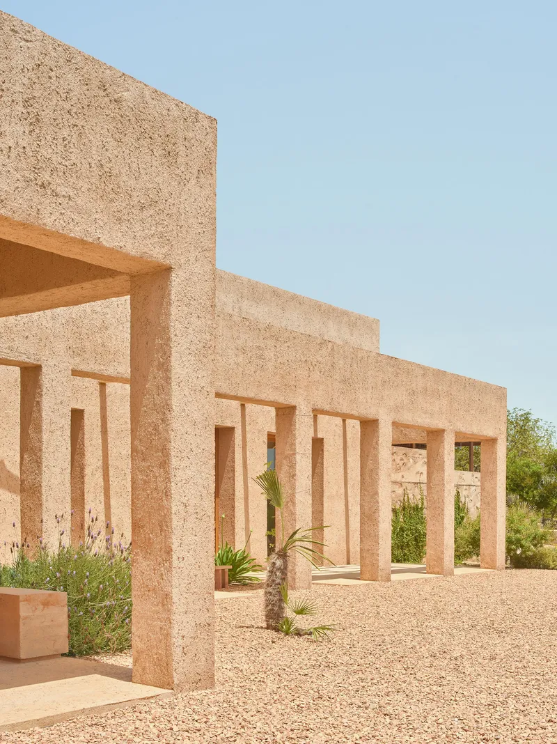 Textured aggregate block colonnade framing a gravel courtyard with planted palms under clear sky