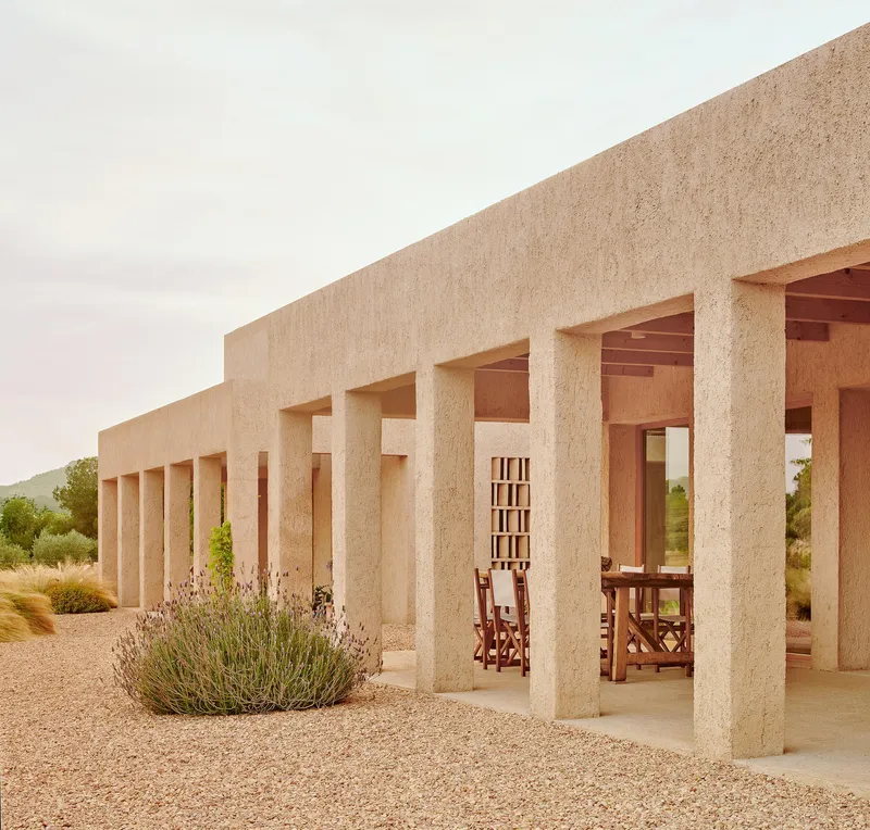 Long colonnade with timber ceiling panels and wooden furniture visible beyond planted grasses
