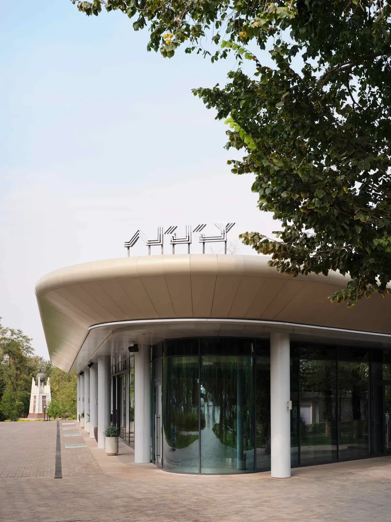 Curved facade with white columns and glazing beneath an overhanging roof framed by tree canopy