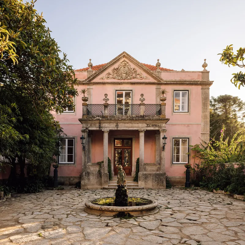Pink stucco facade with pediment and central balcony above a stone fountain in a cobbled courtyard at dusk