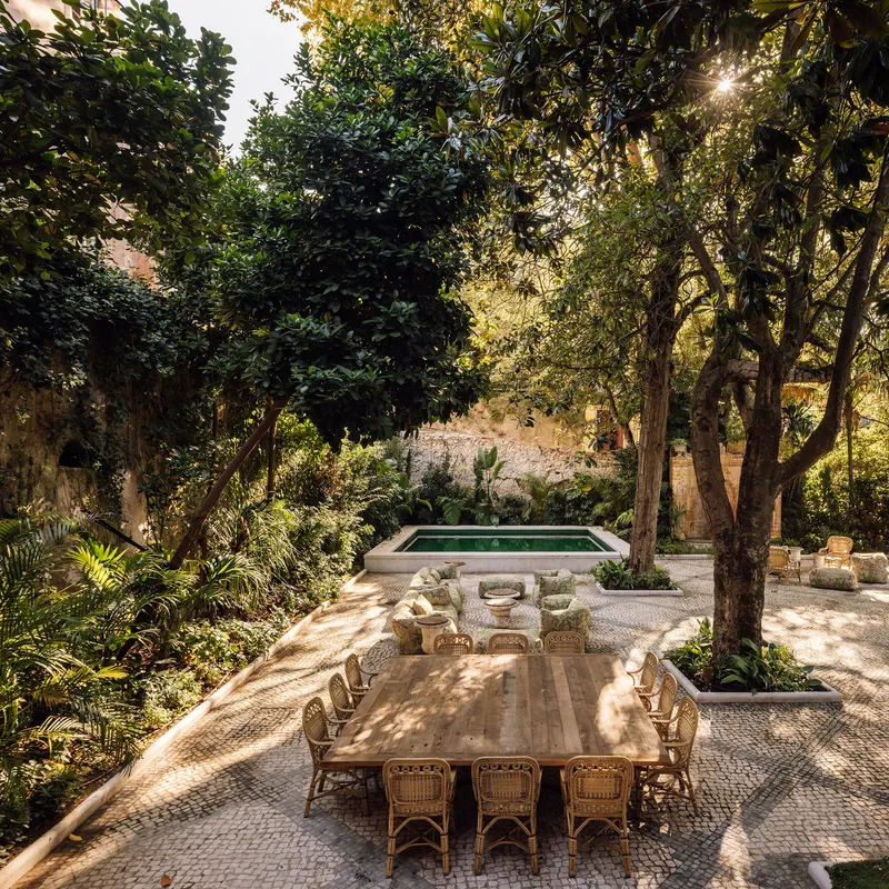 Outdoor terrace with long timber dining table and rectangular pool under dappled sunlight through trees