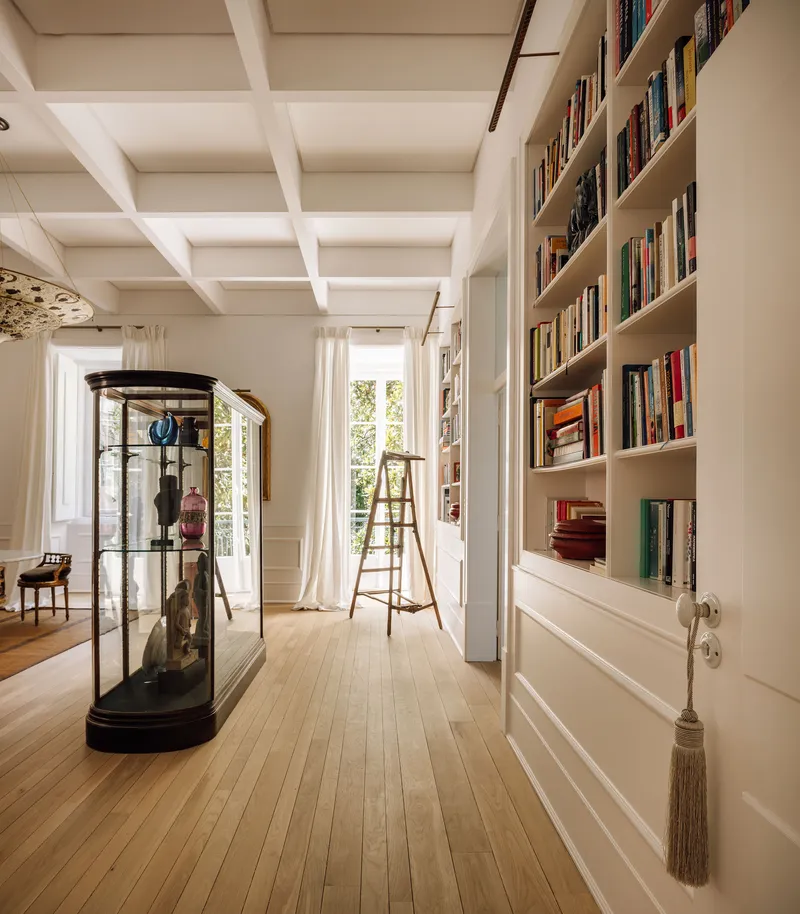 Corridor with coffered ceiling, built-in bookshelves, glass vitrine, and wooden ladder beside tall windows