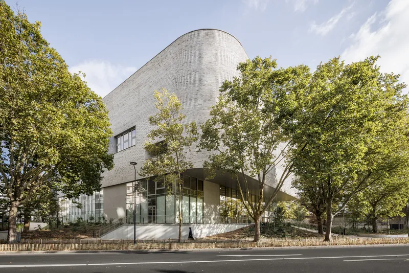 Street view of the curved brick volume rising above a glazed ground floor framed by trees