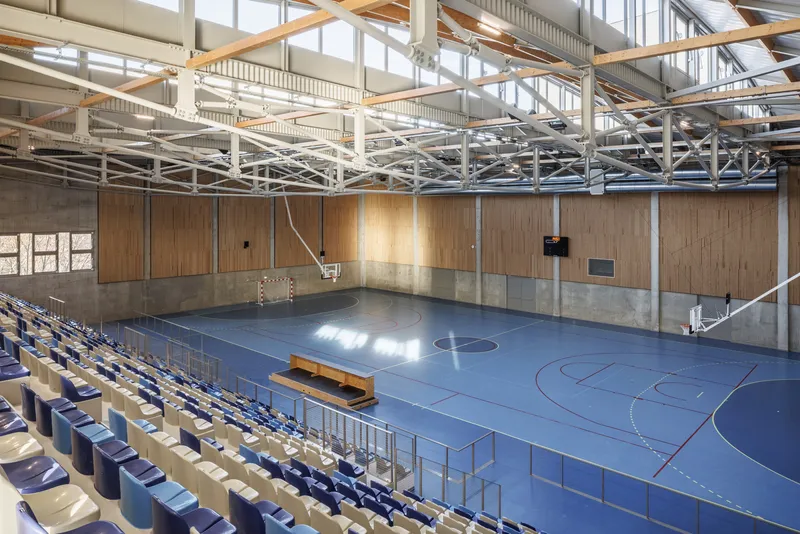 Interior view of the sports hall showing exposed timber and steel trusses above blue flooring and bleachers