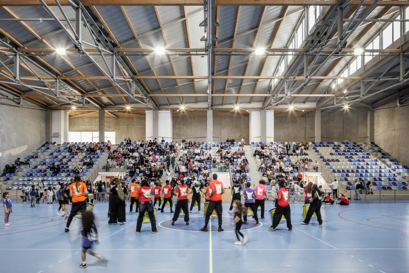 Gymnasium interior filled with people in colored vests beneath the exposed structural ceiling and concrete walls