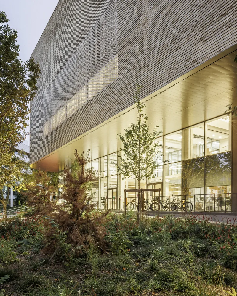 Illuminated glass entrance wall beneath the angled textured facade at dusk with planted trees and parked bicycles