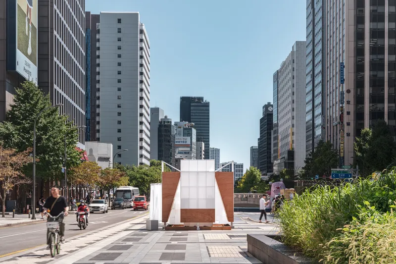 White and wood-paneled pavilion centered in a tree-lined urban plaza with cyclists passing by