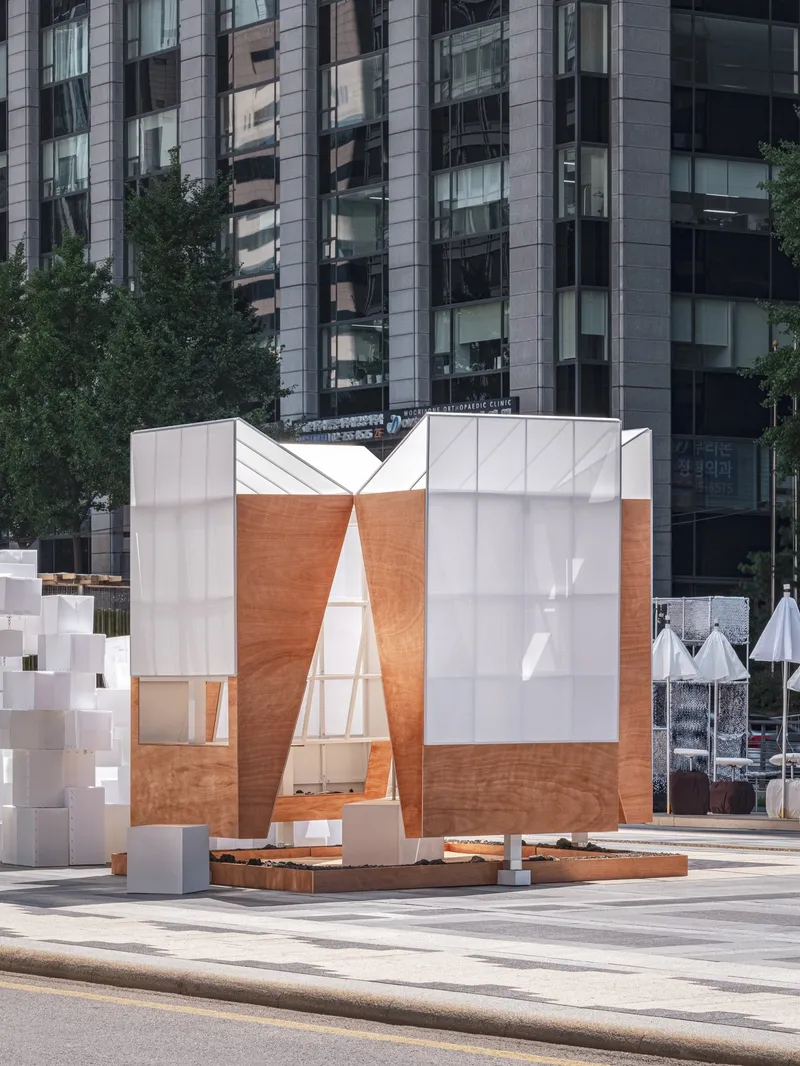 Timber and translucent panel pavilion with triangular openings framing views of residential towers behind
