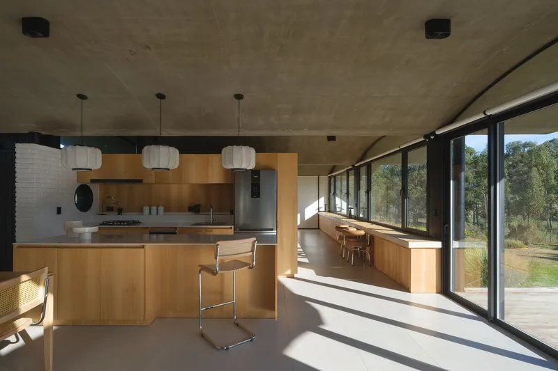 Open-plan kitchen and dining area with exposed concrete ceiling vault and floor-to-ceiling glazing overlooking the landscape