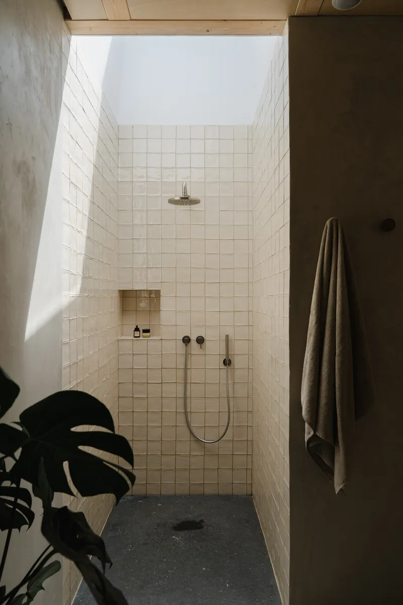 White tiled shower enclosure with skylight above and monstera plant silhouette in foreground