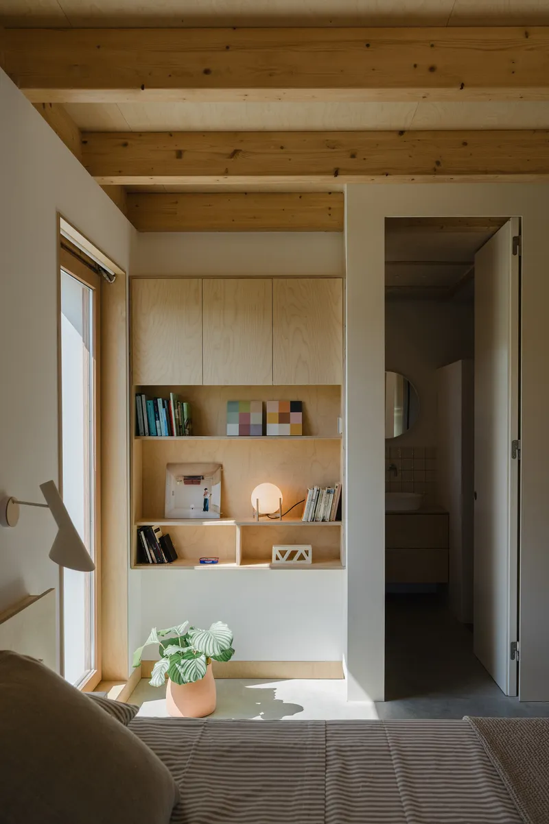 Bedroom with built-in plywood shelving niche beside doorway opening and window casting afternoon light