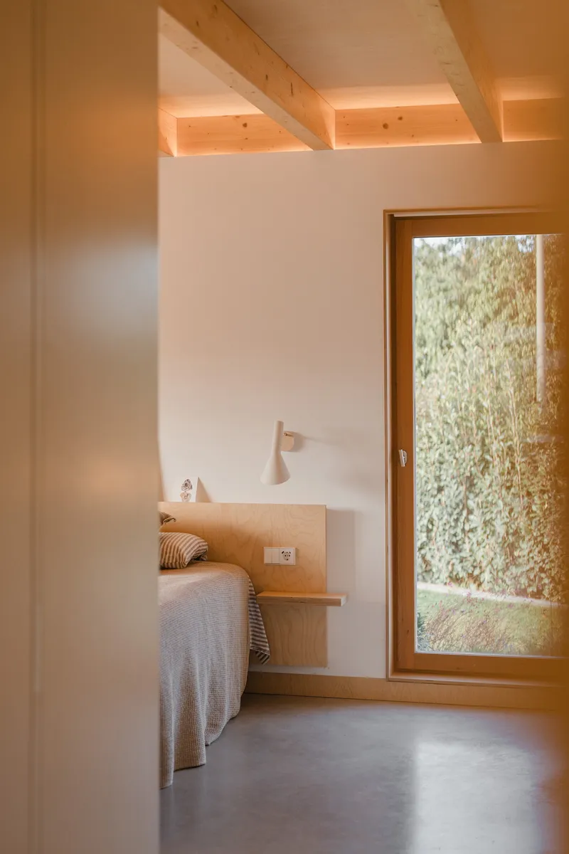 Bedroom with plywood bed frame and timber-beamed ceiling lit by afternoon sunlight through a garden-facing window