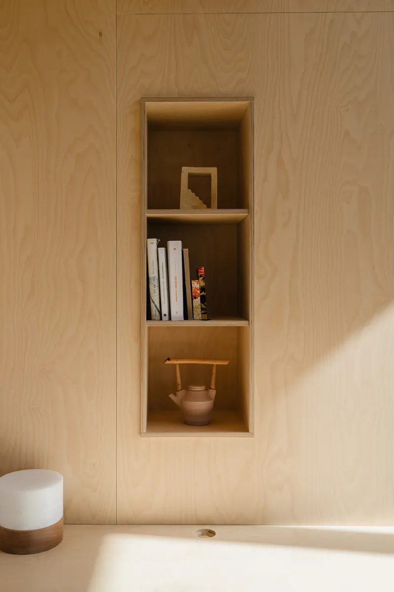 Built-in plywood shelving niche holding books and ceramics within a pale wood wall panel