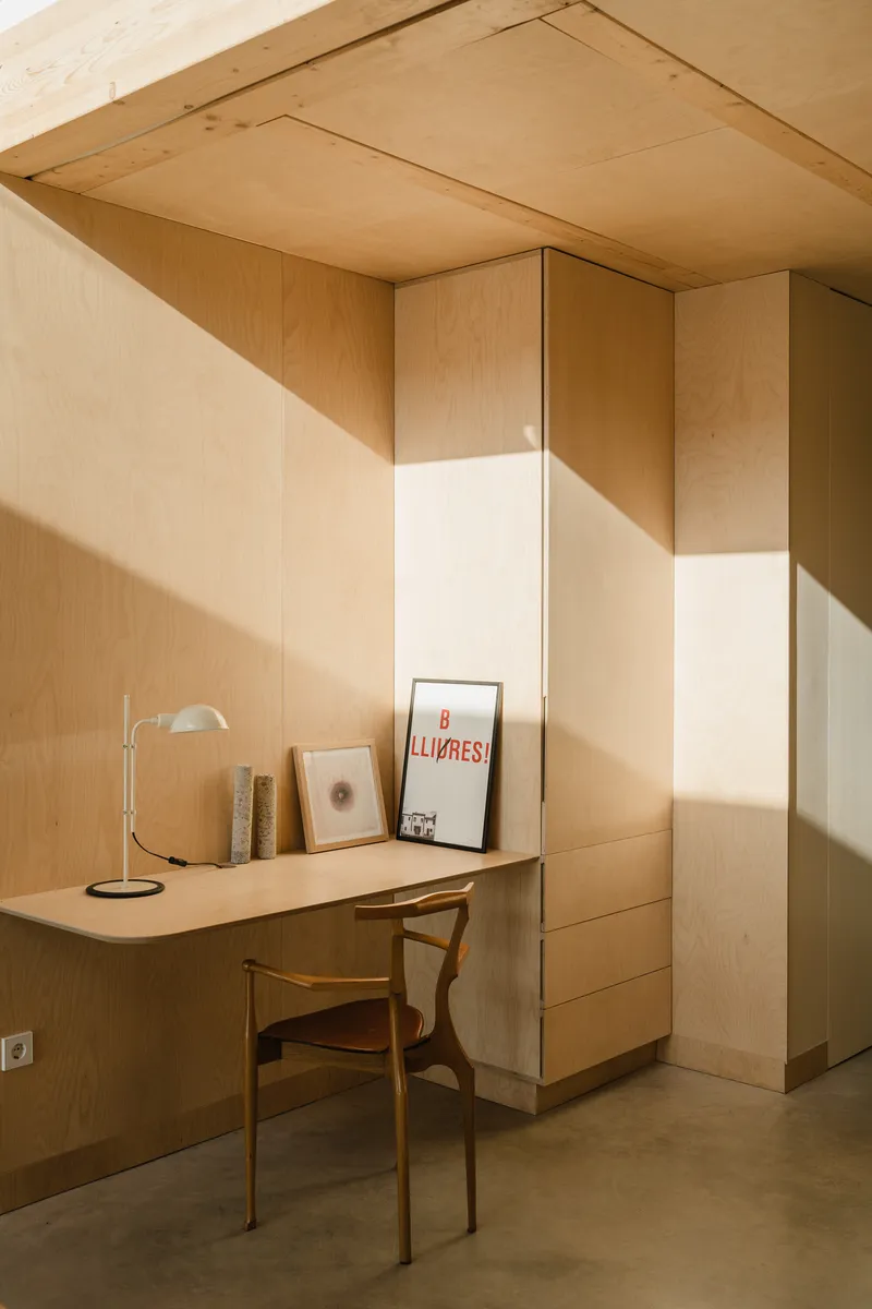 Corner desk with plywood cabinetry and exposed timber ceiling under warm diagonal sunlight