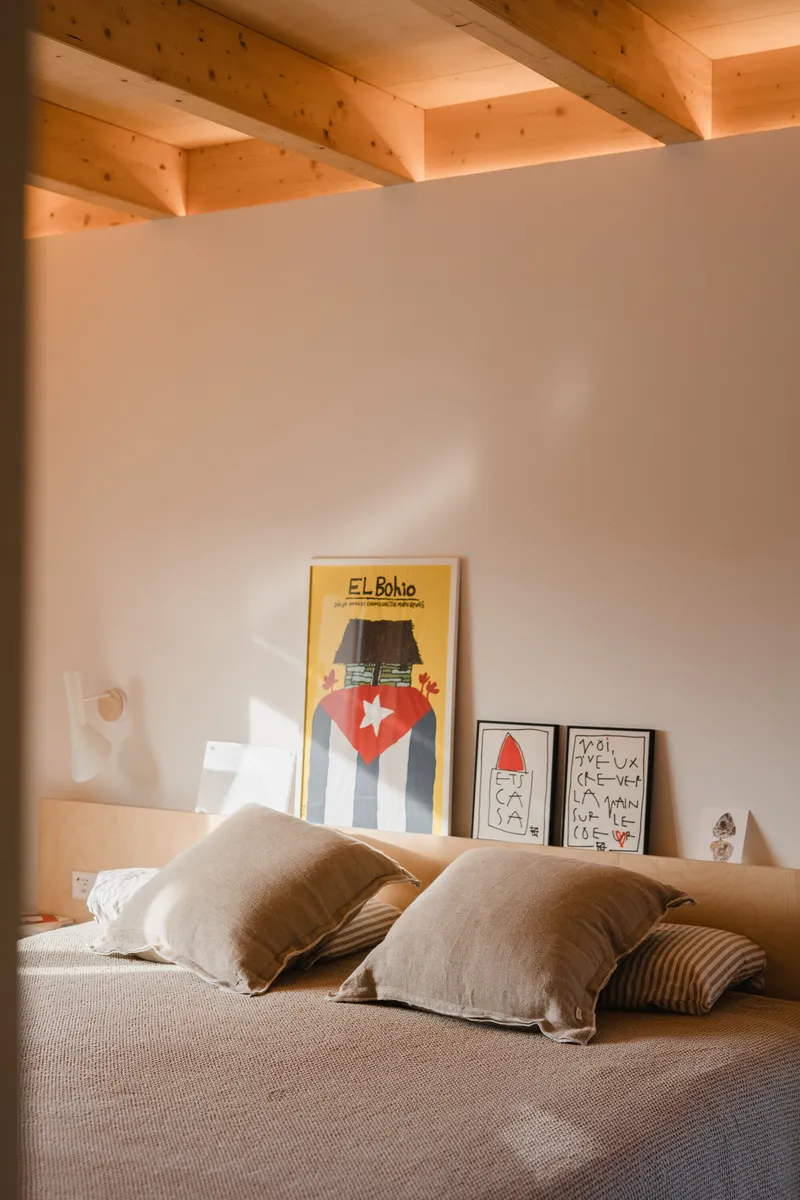 Bedroom headboard wall with framed posters beneath exposed timber joists and indirect cove lighting