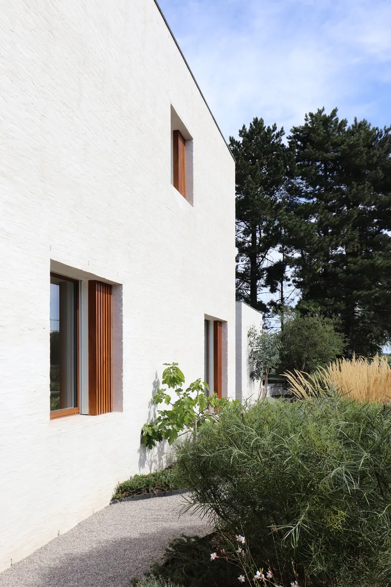 White brick facade with vertical timber shutters flanking deep-set window openings and planted beds