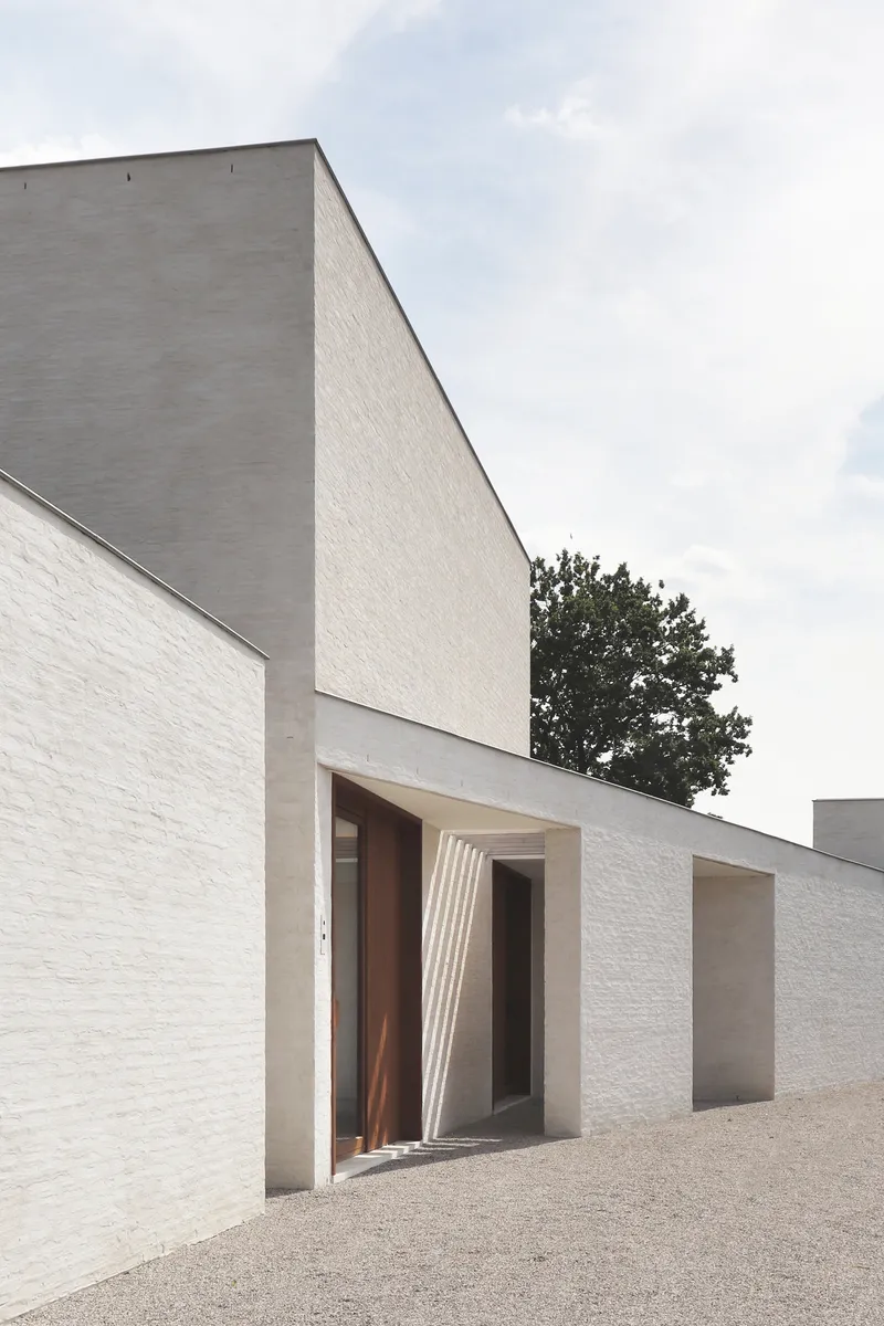Entrance with ribbed concrete canopy framing a timber door set in white brick walls beneath an overcast sky