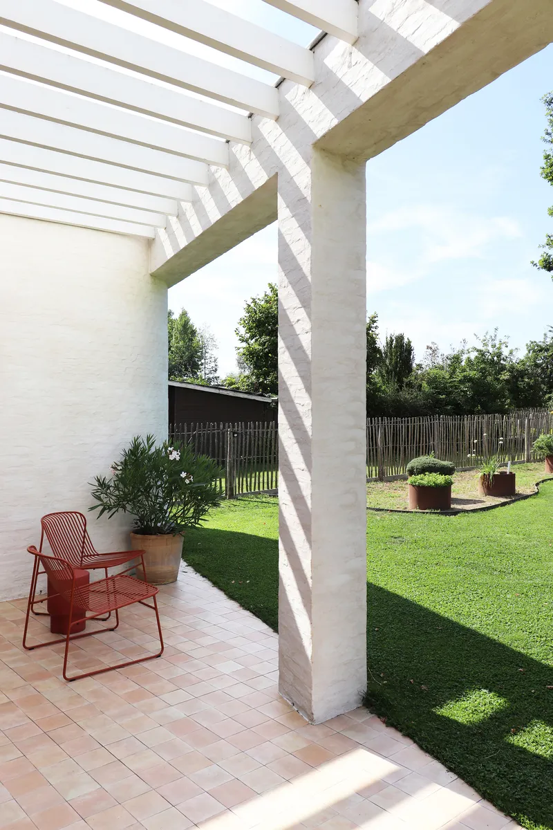 Pergola structure with white slat roof and concrete column casting striped shadows on terracotta paving