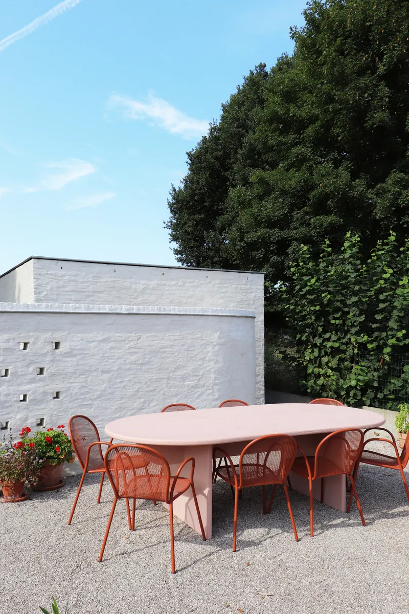 Outdoor gravel terrace with pink oval table and red chairs against white brick wall