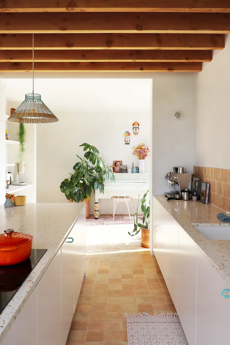 Galley kitchen with terrazzo counters and terracotta floor tile under exposed timber beams looking toward a planted workspace