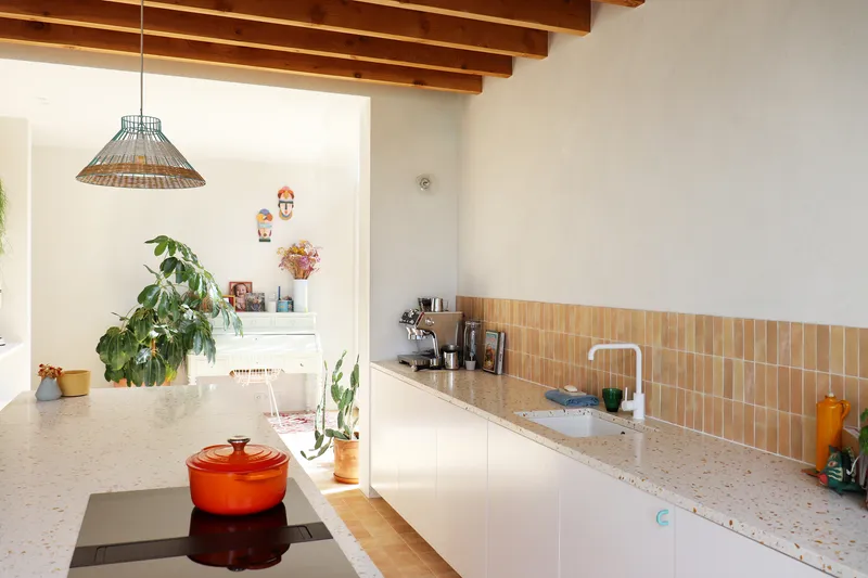 Kitchen with exposed timber beams, white cabinets, terrazzo countertop and terracotta tile backsplash under a woven pendant light